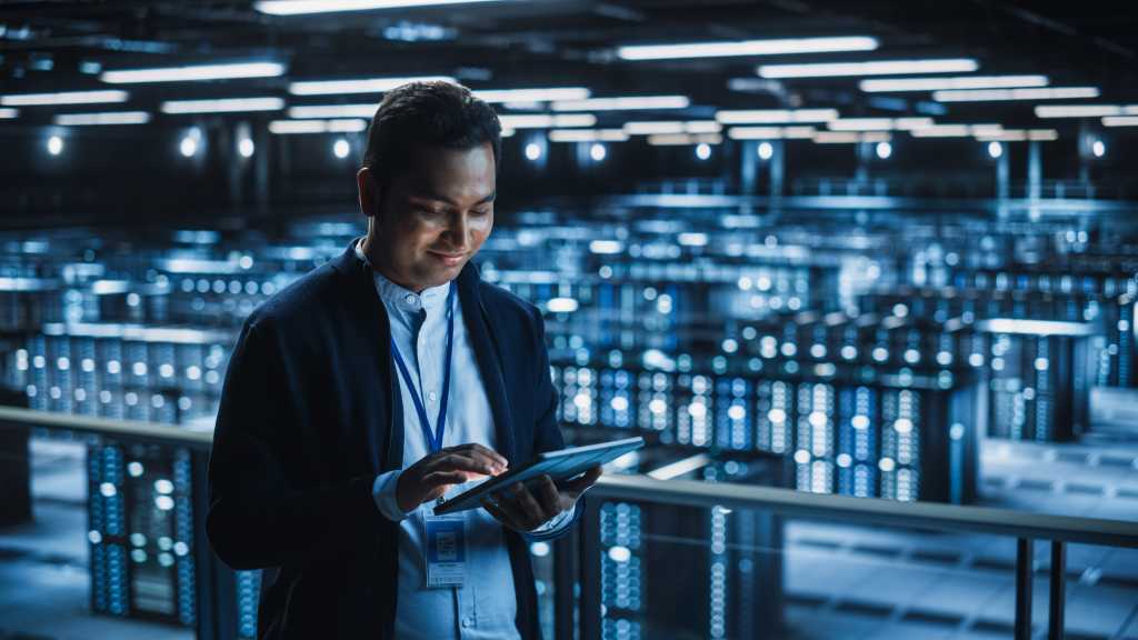 Handsome Smiling IT Specialist Using Tablet Computer in Data Center. Succesful Businessman and e-Business Entrepreneur Overlooking Server Farm Cloud Computing Facility.