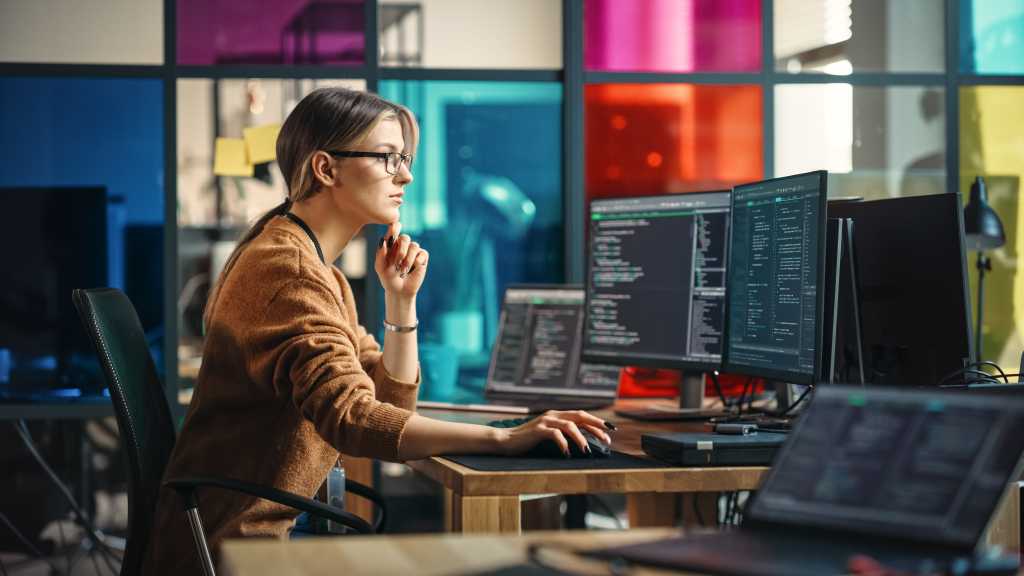 Female Junior Software Engineer Writes Code on Desktop Computer With Two Monitors and Laptop Aside In Stylish Office. Caucasian Woman Working On Artificial Intelligence Service For Big Tech Company.