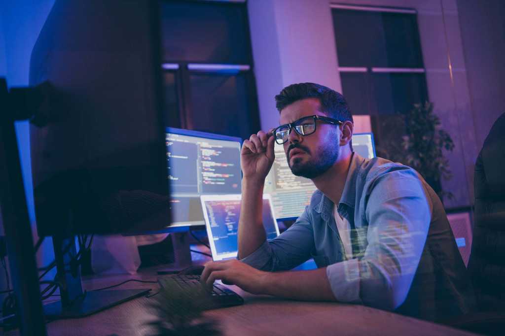 Profile photo of a developer / programmer reviewing code on monitors in his workspace.