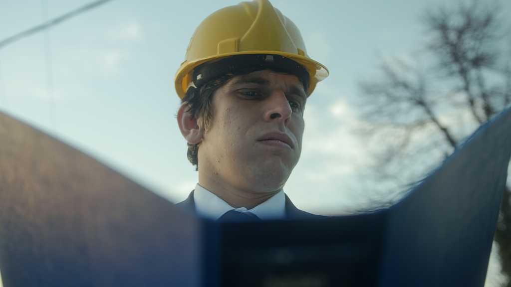 Beardless Construction Engineer or Contractor in a Helmet and Suit Examining Blueprints in a Blue Folder and Getting Frustrated over a Faulty Detail